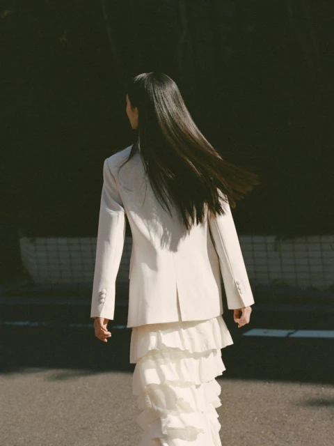 Back view of a young woman with long black hair walking on a city street wearing a white blazer and tiered skirt, bathed in sunlight.