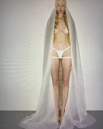 Model wearing a sheer floor-length white veil cape and matching lingerie set against a neutral grey studio background