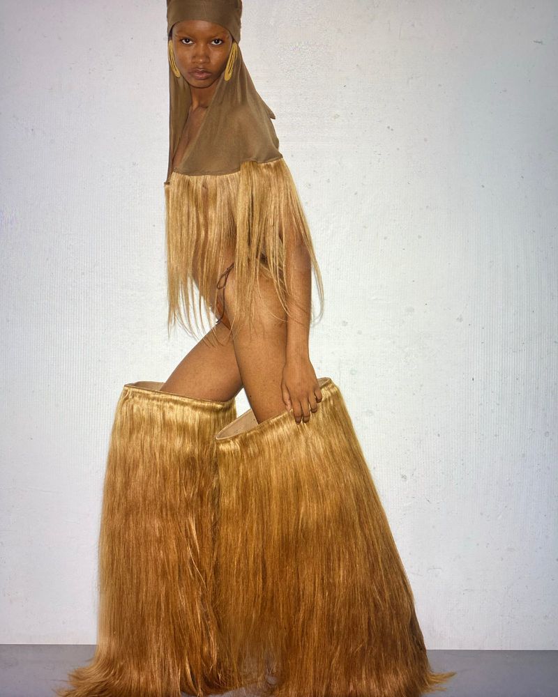 Striking studio portrait of a Black woman wearing an elaborate fringed costume and head covering, posing against a plain grey background.