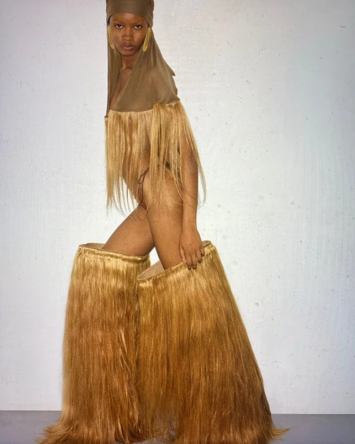 Striking studio portrait of a Black woman wearing an elaborate fringed costume and head covering, posing against a plain grey background.