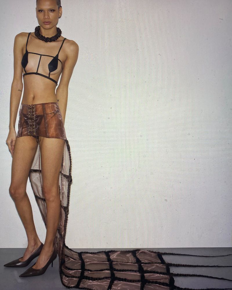Full-length fashion shot of a woman with short hair wearing a sheer brown skirt with a long train, heels, and a structured top against a white backdrop.