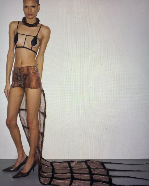 Full-length fashion shot of a woman with short hair wearing a sheer brown skirt with a long train, heels, and a structured top against a white backdrop.