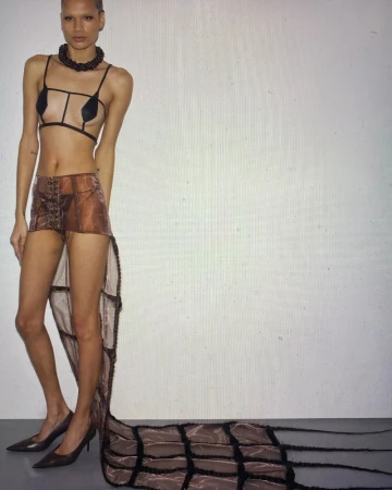 Full-length fashion shot of a woman with short hair wearing a sheer brown skirt with a long train, heels, and a structured top against a white backdrop.