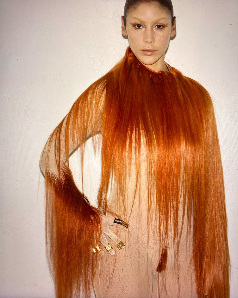 A woman with artistic makeup and long red hair draped as a garment poses indoors with a plain white background for a beauty shot.