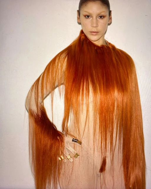 A woman with artistic makeup and long red hair draped as a garment poses indoors with a plain white background for a beauty shot.