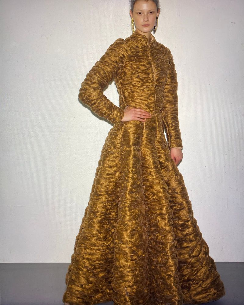 Model standing against a white seamless background wearing a textured golden brown long sleeve dress