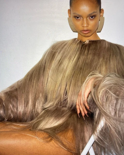 A person with tan skin is posing in a full shot. They are wearing a garment with many strands of blond hair, large earrings, and makeup.