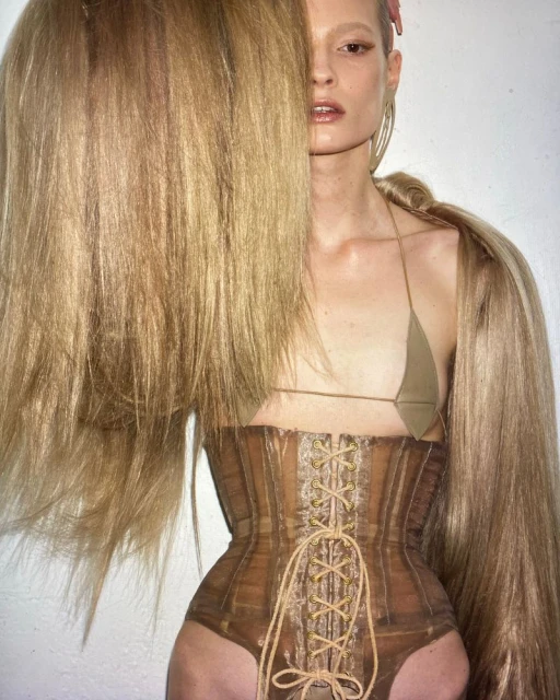 A fair-skinned woman with blonde hair poses in a studio wearing a tan bra top and corset with blonde hair extensions wrapped around her shoulders.