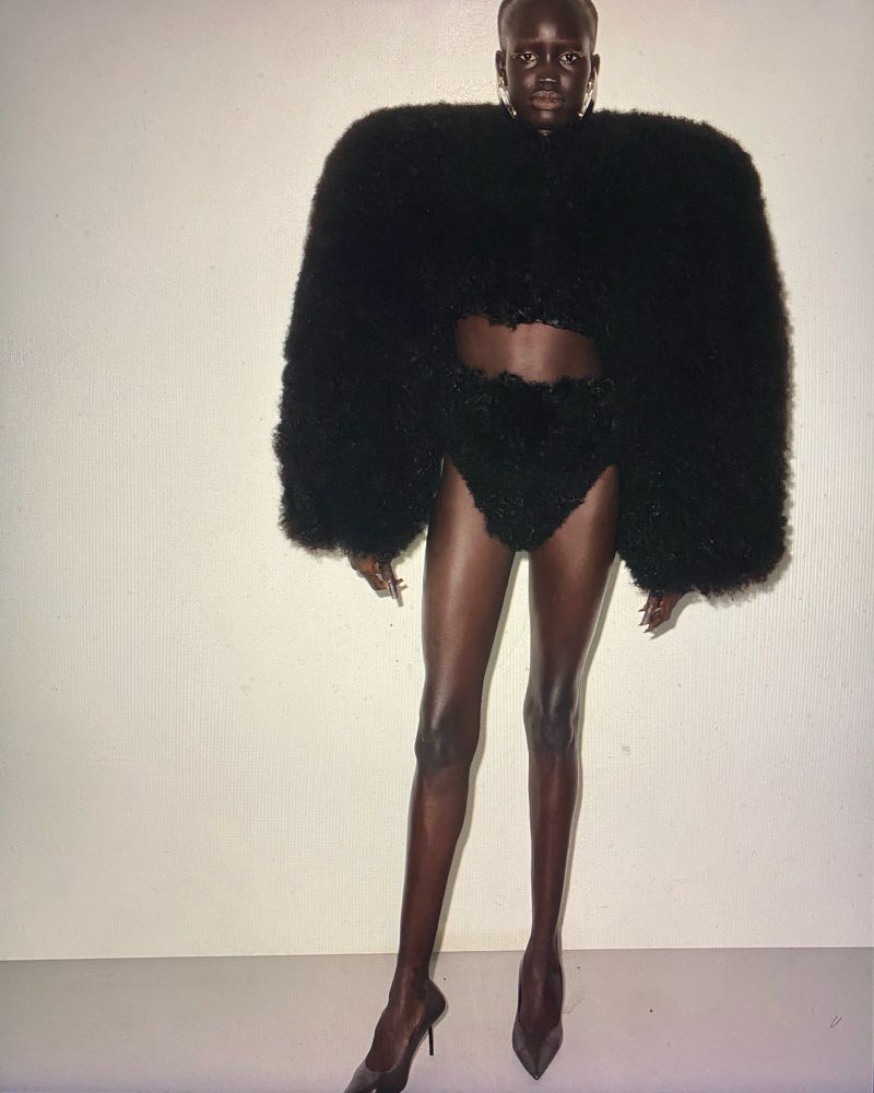 A striking fashion image featuring a black female model wearing avant-garde black high-fashion attire and high heels against a white backdrop.