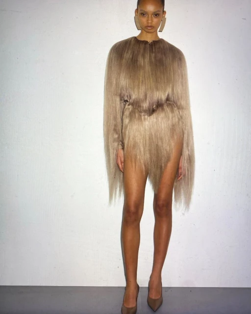 Fashion model poses in a unique beige shaggy dress and high heels against a neutral backdrop, showcasing avant-garde style.