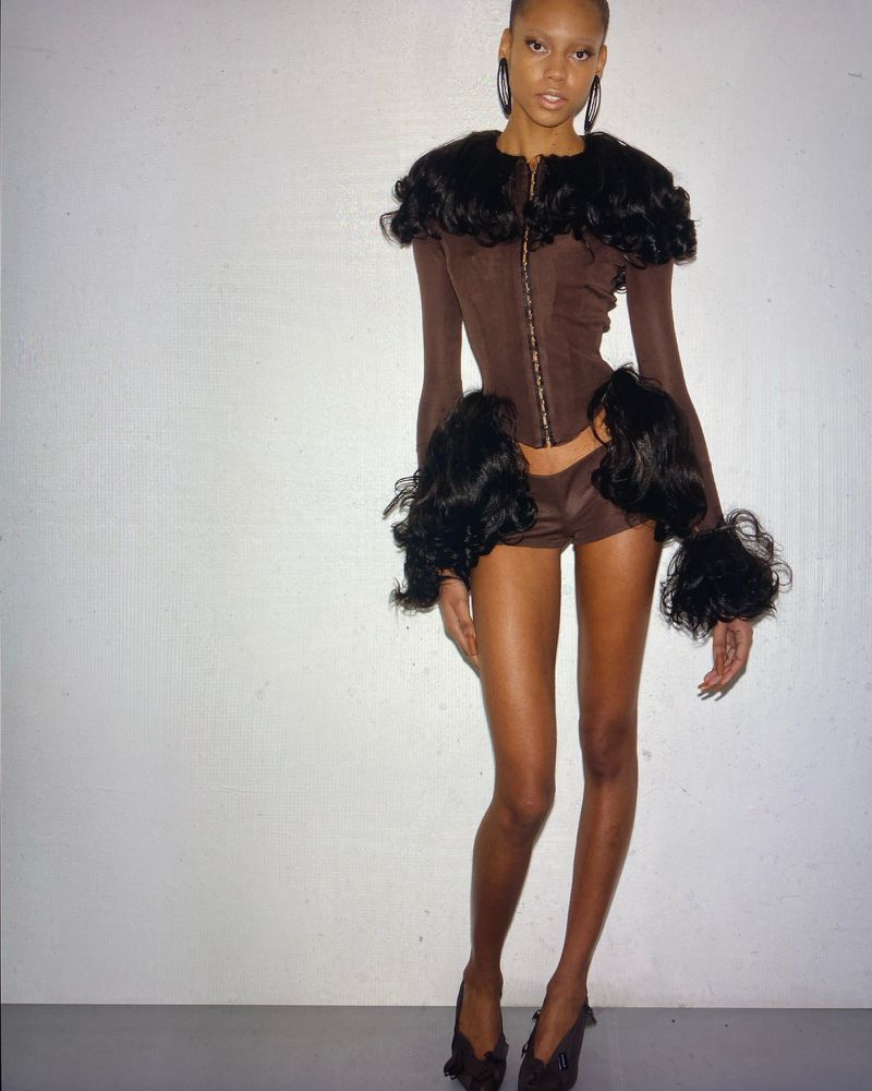 A tanned woman in a brown short set with dark, curly accents, accessorized with large hoop earrings, poses against a white wall.