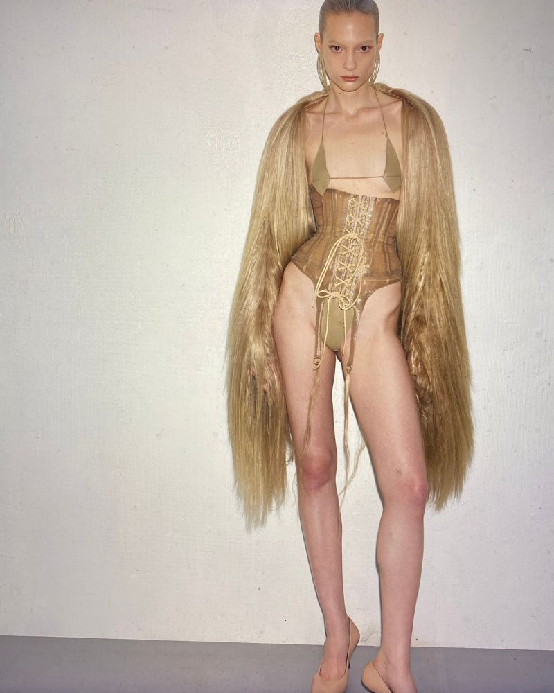 A woman in a corset and fur coat stands in a studio. She is wearing earrings and high heels. The background is a plain white wall.