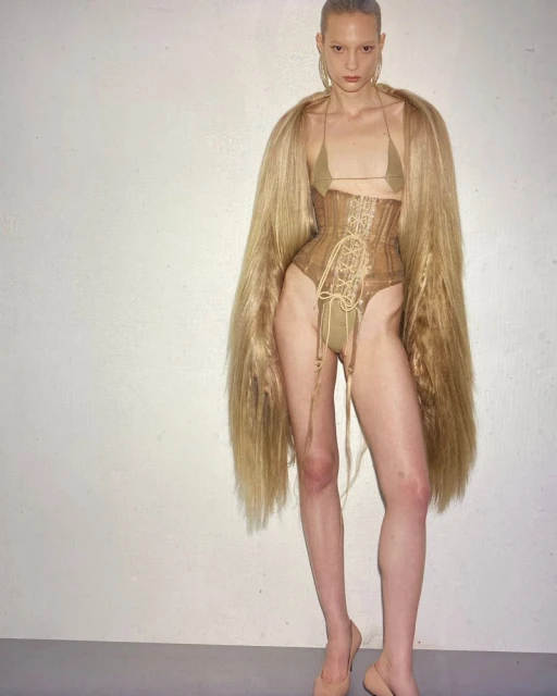 A woman in a corset and fur coat stands in a studio. She is wearing earrings and high heels. The background is a plain white wall.