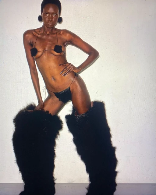 A nude African woman is striking a full pose in black furry boots and decorative pasties in a studio setting. She faces the camera.