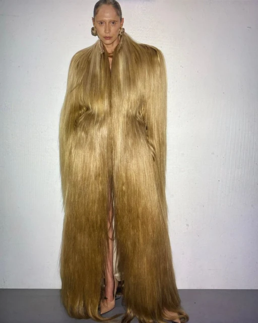 Adult woman posing in a long coat of long hair in front of a white wall. Fashion-forward editorial style, blond hair texture.