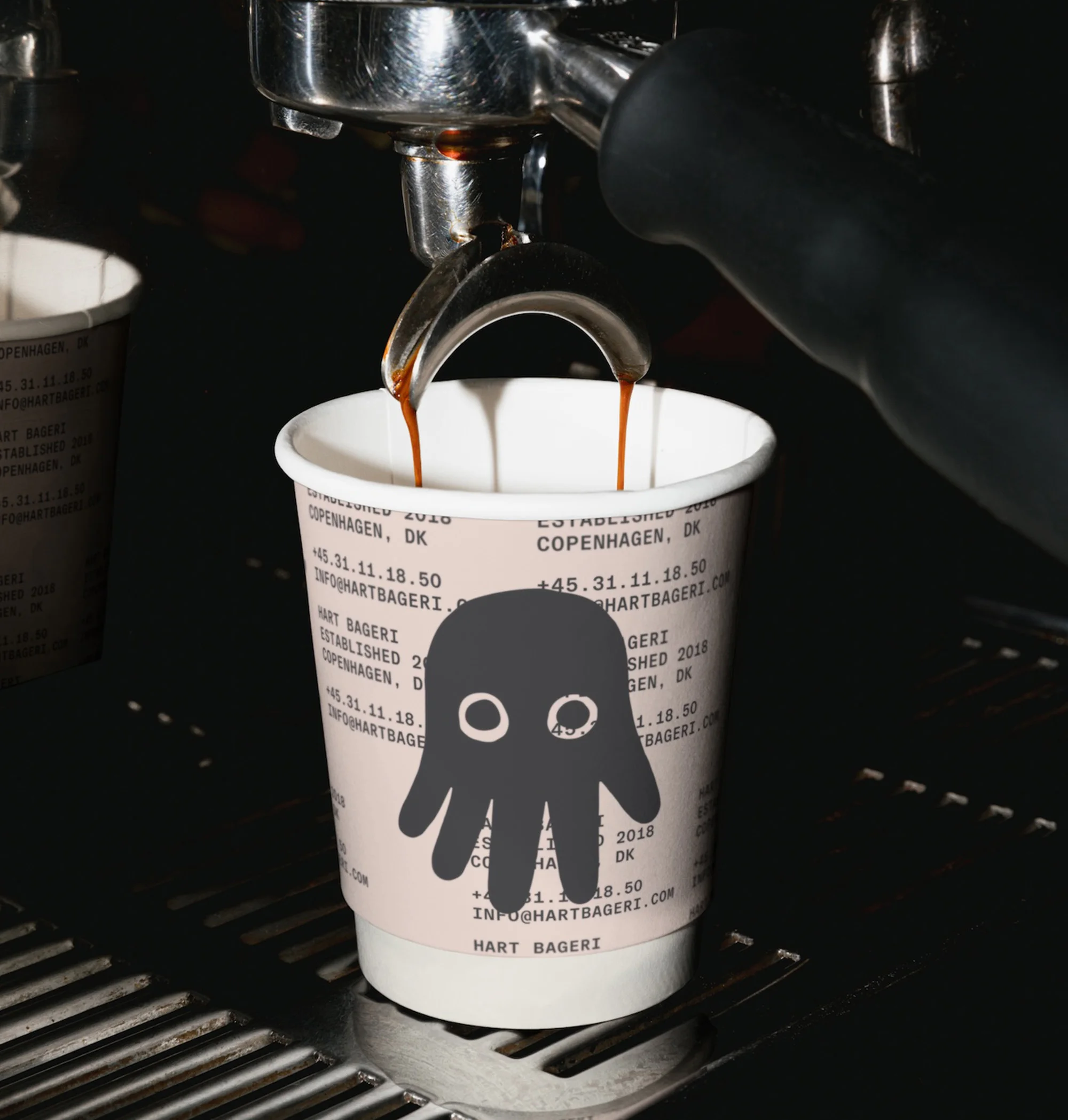 Espresso pours into a paper cup with a cartoon ghost, on an espresso machine. Bakery branding and location text visible on cup.