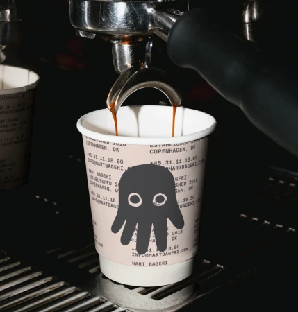 Espresso pours into a paper cup with a cartoon ghost, on an espresso machine. Bakery branding and location text visible on cup.