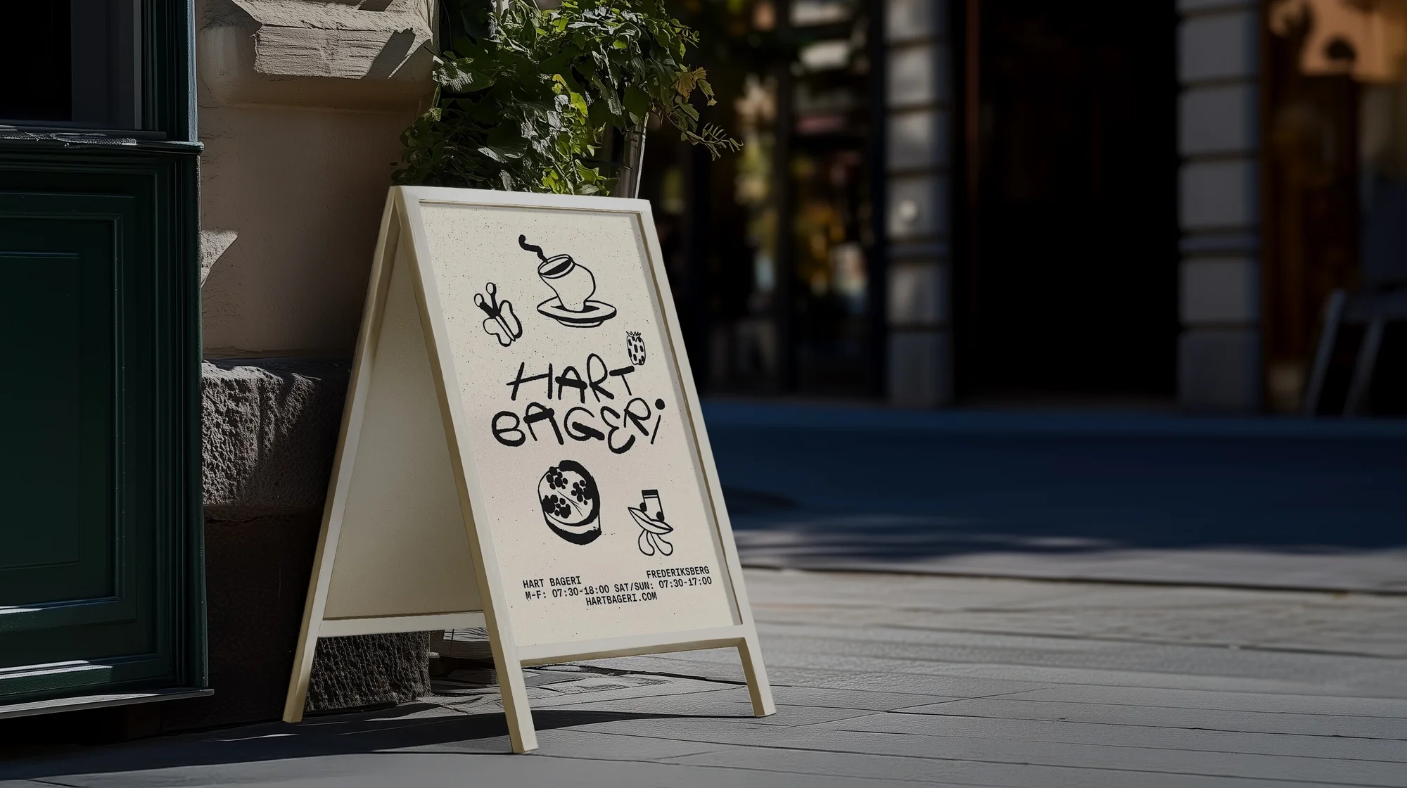 A-frame sign with bakery logo and hours on a sidewalk, with a building facade and greenery as a backdrop on a sunny day.
