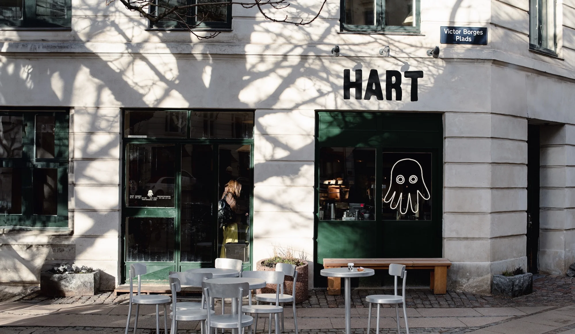 Exterior of Hart Bakery with outdoor seating. Shadow patterns cast by tree branches on the building's facade. Neutral color palette.