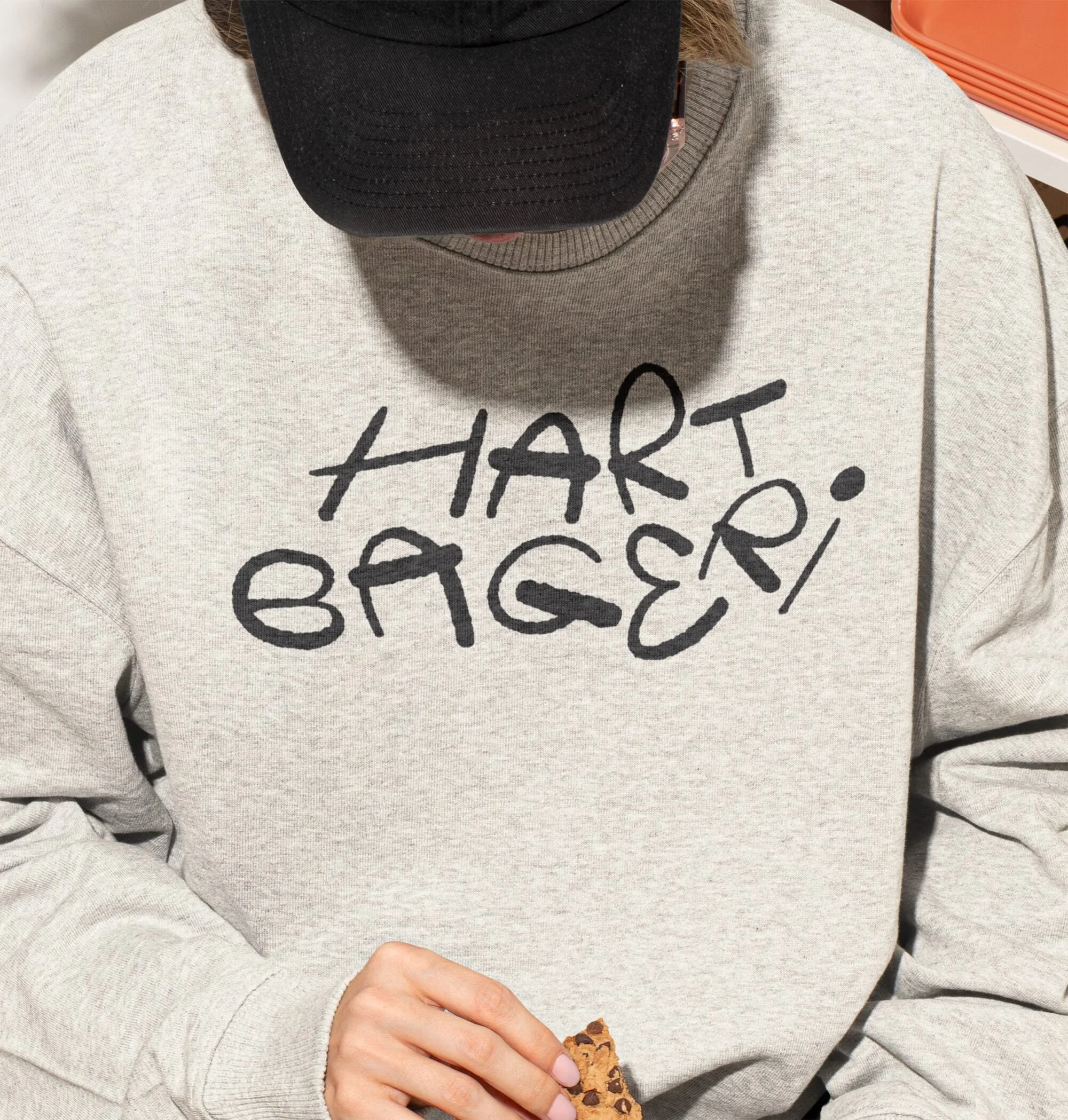 Person wearing a grey sweater with a black logo and a black baseball cap, holding a chocolate chip cookie in their fingers.