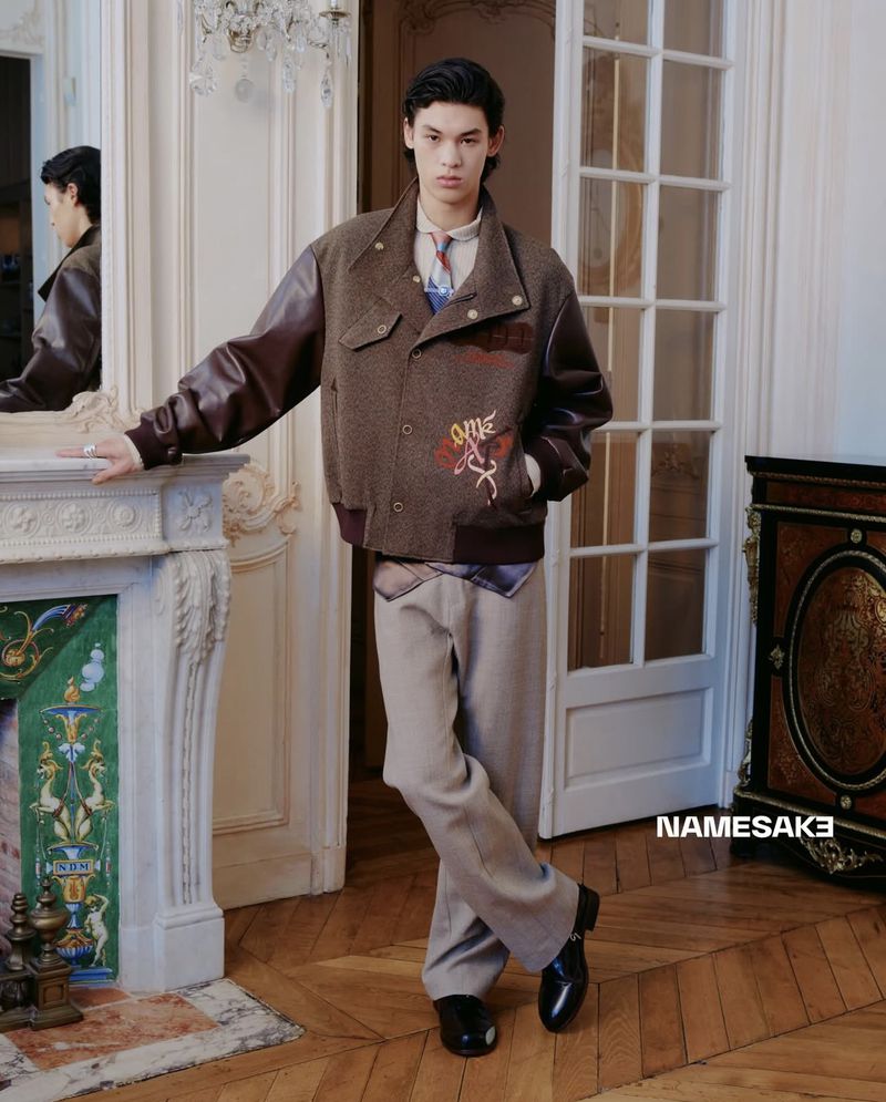 Young man poses in an indoor setting, wearing a stylish brown bomber jacket with leather sleeves and beige trousers. 