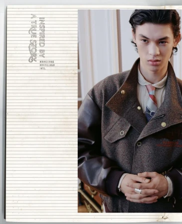 Portrait of a young adult wearing a brown jacket, tie, and ring, shot indoors with a magazine cover featuring lined paper on the left.