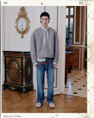 Male fashion model poses in neutral-toned jacket and blue jeans in an ornate room with hardwood floors and antique furniture.