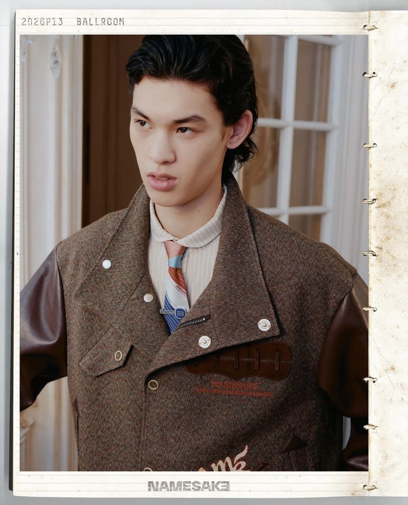 Portrait of a young Asian man in a brown jacket with leather sleeves and patterned tie standing indoors in front of a white door.