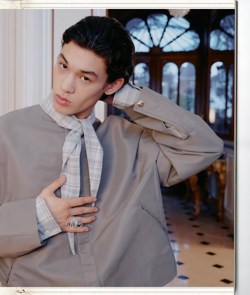 Indoor portrait of a slender young adult male wearing a grey blazer with a patterned scarf, posing with hand on his chest in an interior.