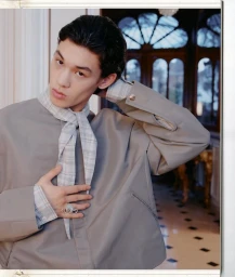 Model wearing a grey jacket over a plaid shirt and scarf, adjusting his hair in a classic interior setting for NAMESAKE F/W 26