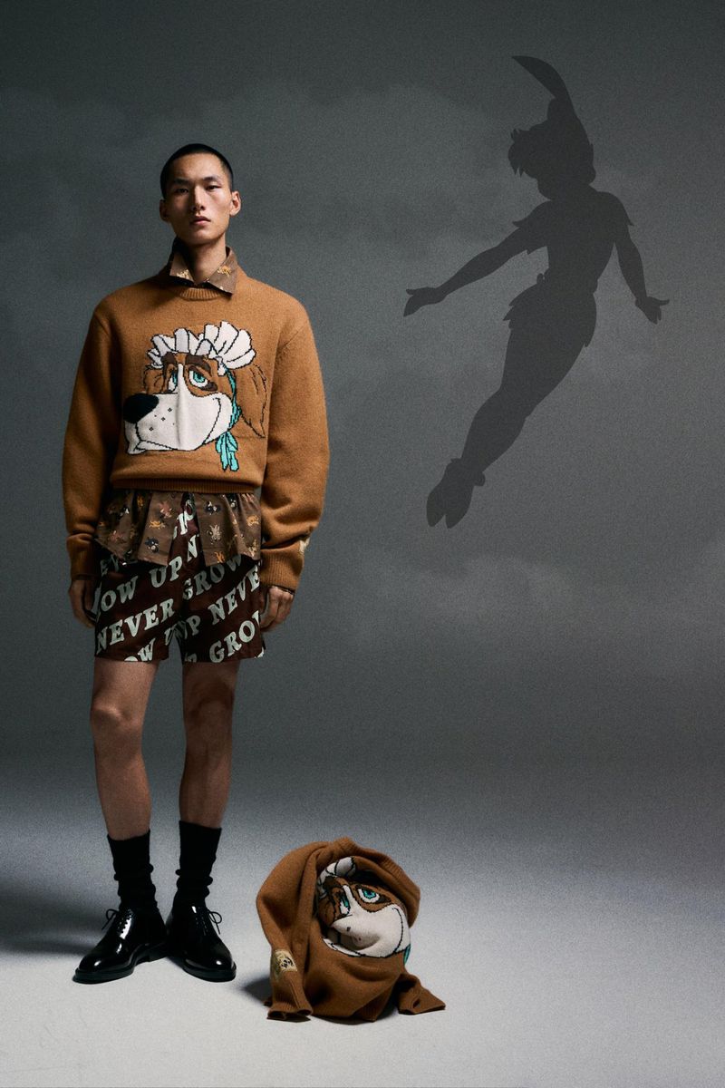 Full-length studio shot of a slender Asian male model wearing a cartoon dog sweater, shorts, black socks, dress shoes, and a Peter Pan shadow.
