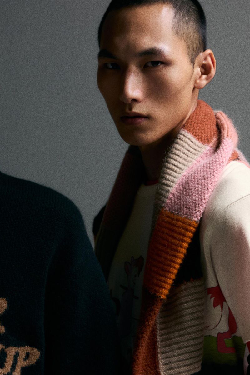 Close-up studio shot of a fashionable young Asian man with a buzzcut, wearing a colorful knitted scarf and a printed sweatshirt.