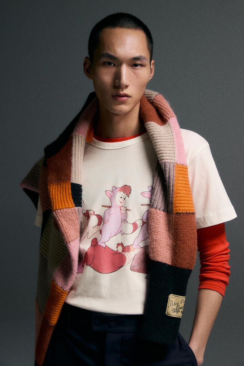 Asian male model poses in a photo studio, wearing a graphic T-shirt and a knitted patchwork scarf, against a grey backdrop.