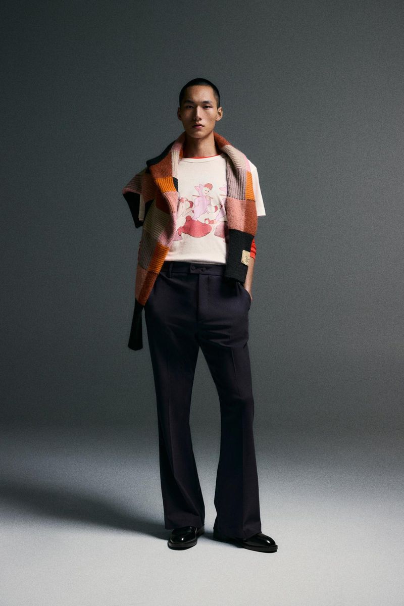 Adult male poses indoors wearing a white t-shirt with a graphic, dark trousers, black shoes, and a multi-colored, knit scarf.