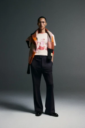 Model wearing a printed t-shirt and dark trousers with a patterned scarf draped over shoulders against a grey studio background.