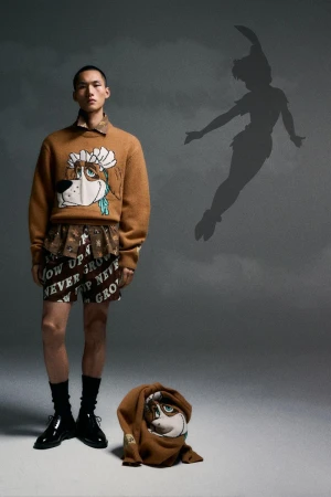 Model wearing a brown Disney character sweater and patterned shorts standing against a grey studio backdrop with a Peter Pan shadow