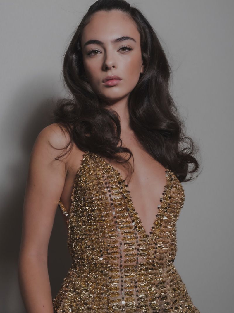 Portrait of a young adult woman with long brown hair wearing a gold beaded dress in front of a solid grey background.