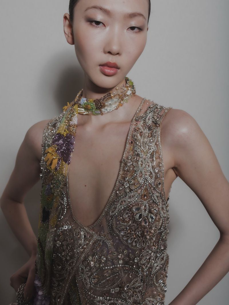 Elegant Asian woman in a sleeveless embellished gown with a floral scarf, posing indoors against a plain light background.