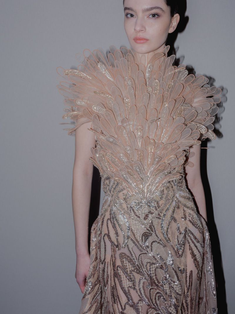 A slender woman poses in a feathered dress, illuminated by soft lighting against a neutral backdrop, showcasing fashion and style.
