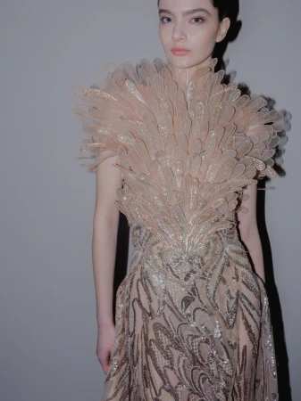 A slender woman poses in a feathered dress, illuminated by soft lighting against a neutral backdrop, showcasing fashion and style.