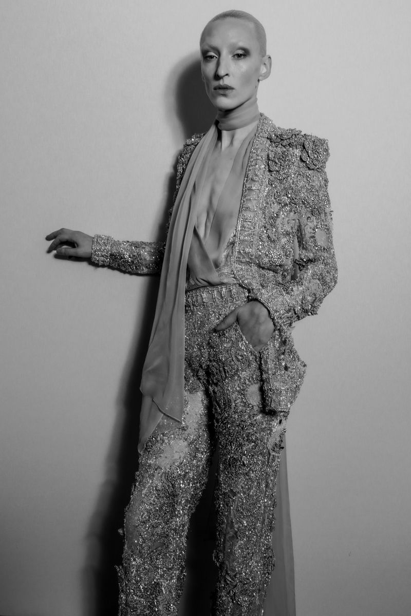 Androgynous fashion model poses in tailored jacket and pants covered in shimmering embellishments and draped chiffon scarf in studio.