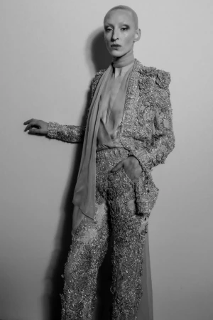 Androgynous fashion model poses in tailored jacket and pants covered in shimmering embellishments and draped chiffon scarf in studio.