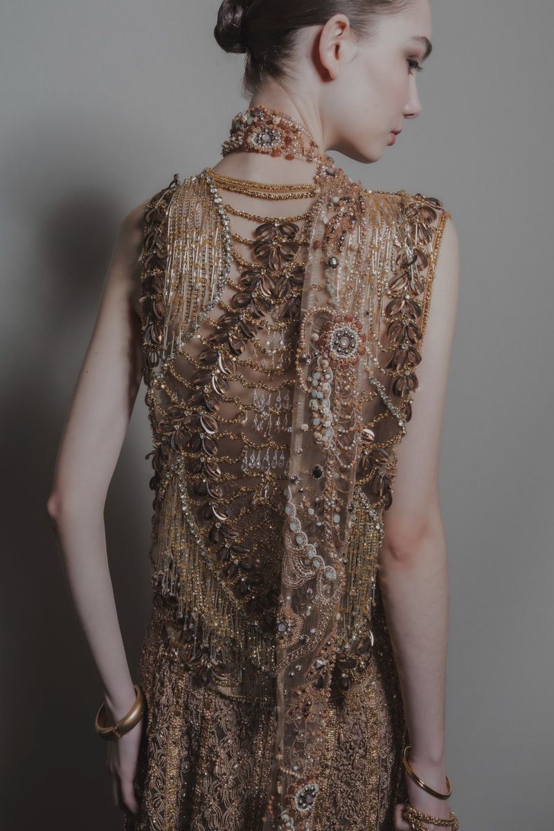 Woman wearing a golden, jeweled gown and gold jewelry from the back, revealing the intricate gold pattern on the dress and shawl.