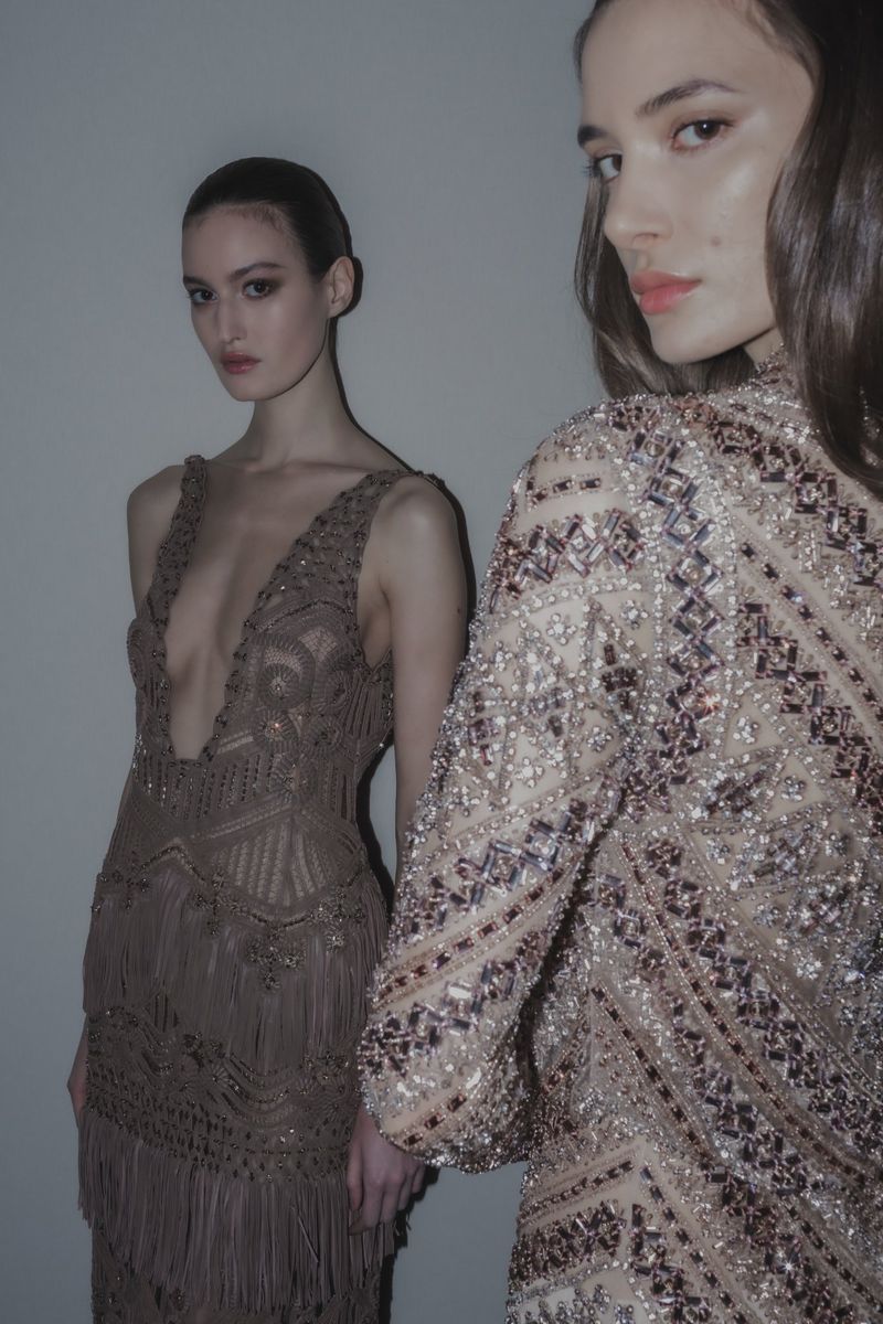 Two fashion models pose indoors wearing intricately beaded gowns in a studio setting with neutral background and diffused lighting.
