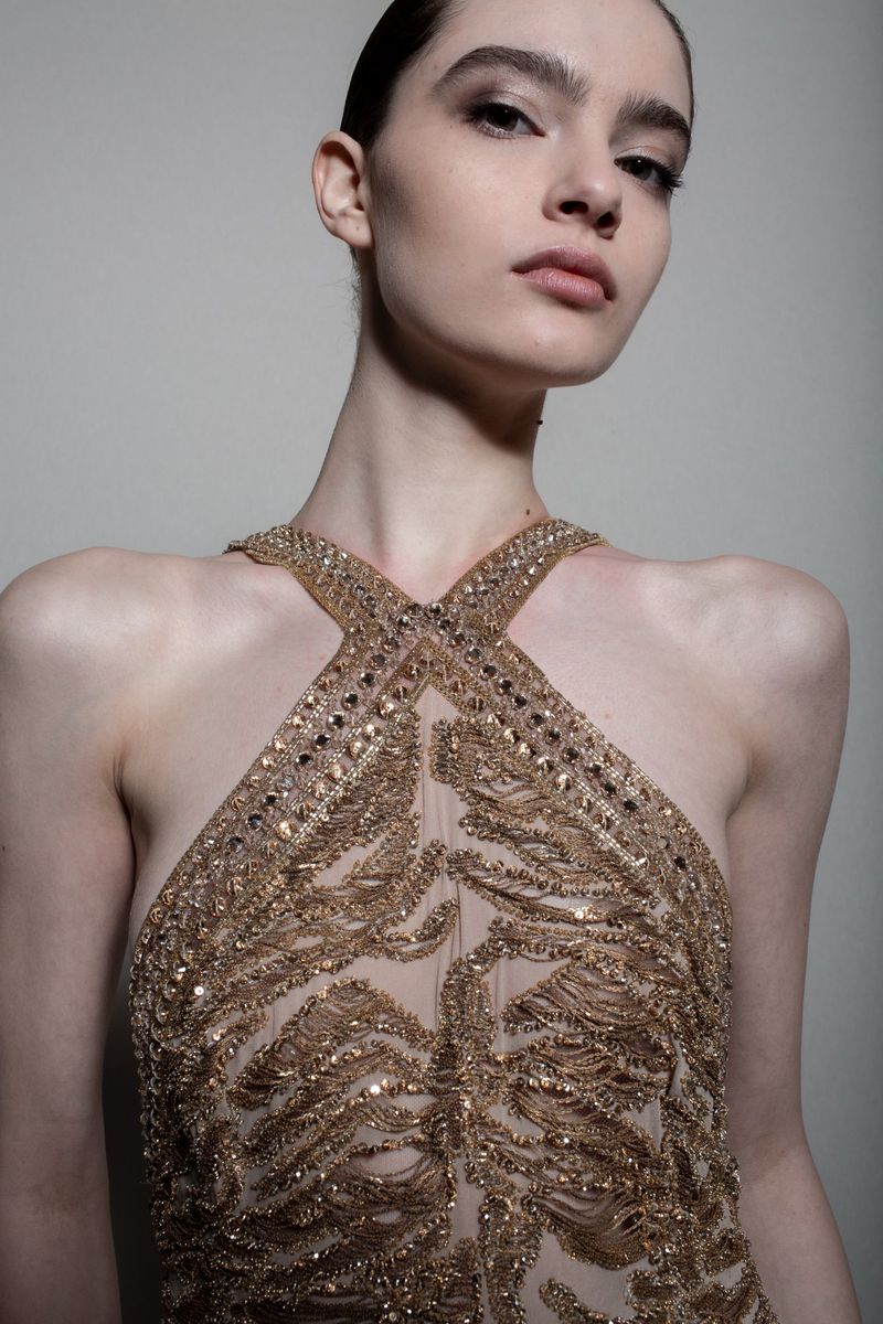 Close-up of a young woman in a beaded halter dress, posing against a neutral background. Beauty shot emphasizes her makeup and attire.
