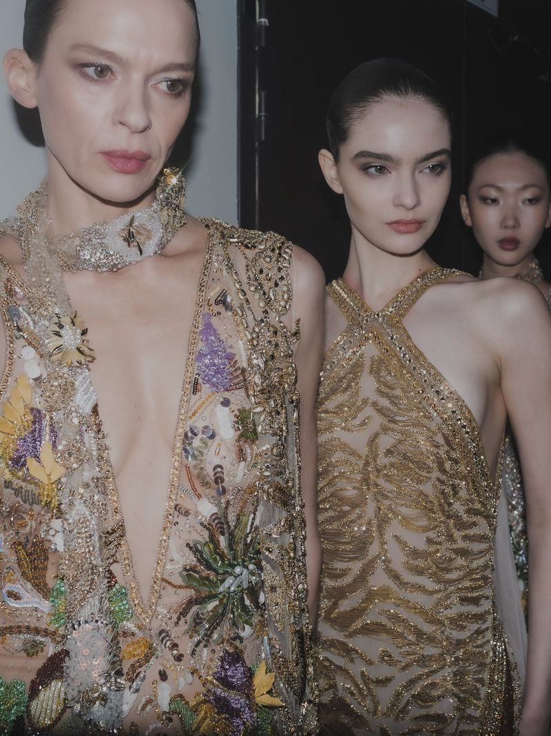 Three runway models backstage wear embellished, semi-sheer gowns in a dimly lit indoor setting.