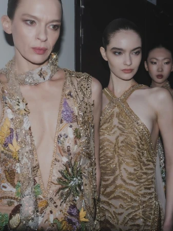 Three runway models backstage wear embellished, semi-sheer gowns in a dimly lit indoor setting.