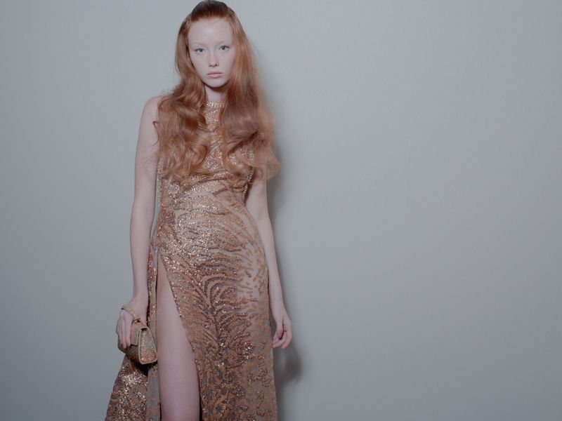 A red-haired fashion model wears a floor-length, sequined gown with a high slit while posing in front of a solid grey background indoors.