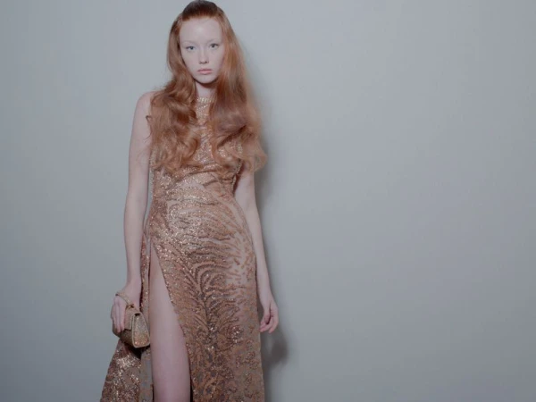 A red-haired fashion model wears a floor-length, sequined gown with a high slit while posing in front of a solid grey background indoors.