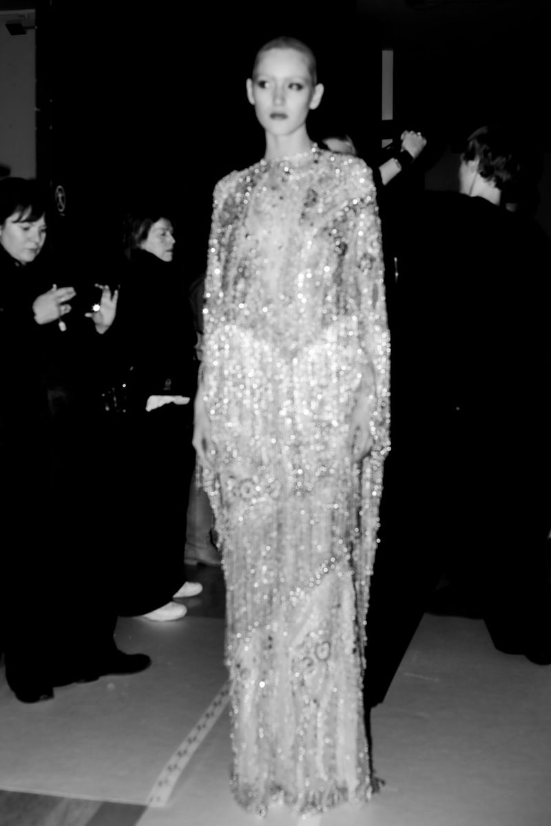 Model wearing beaded gown on fashion runway, surrounded by onlookers in a monochromatic photo capturing a backstage scene.